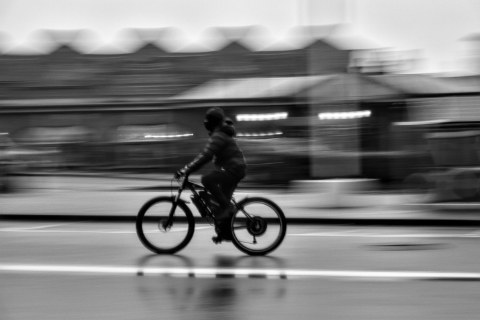 Black and white panning photograph of a cyclist speeding along a street, heavily blurring the background buildings.