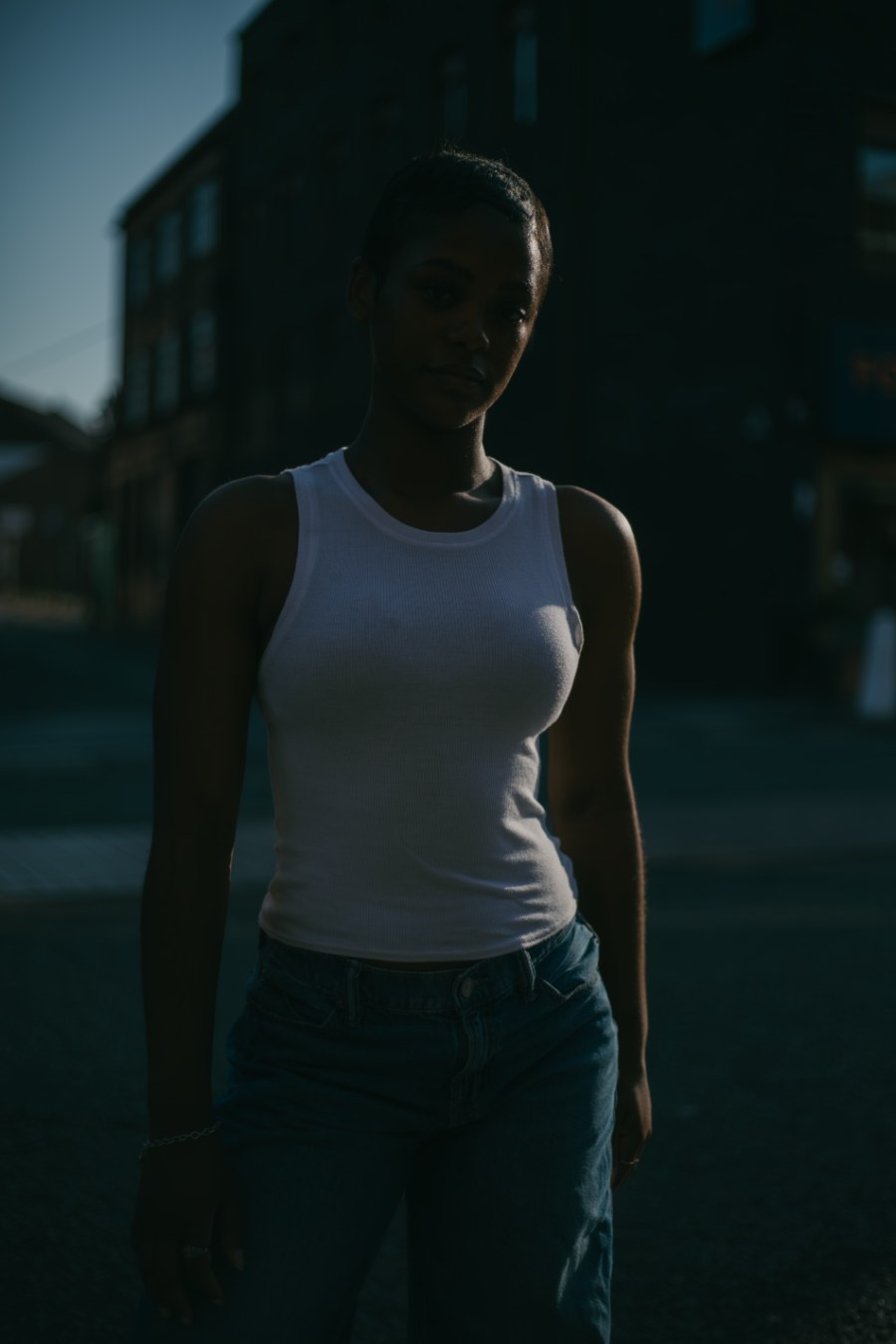 A woman in a white tank top and jeans stands in deep, moody shadow on a quiet street.