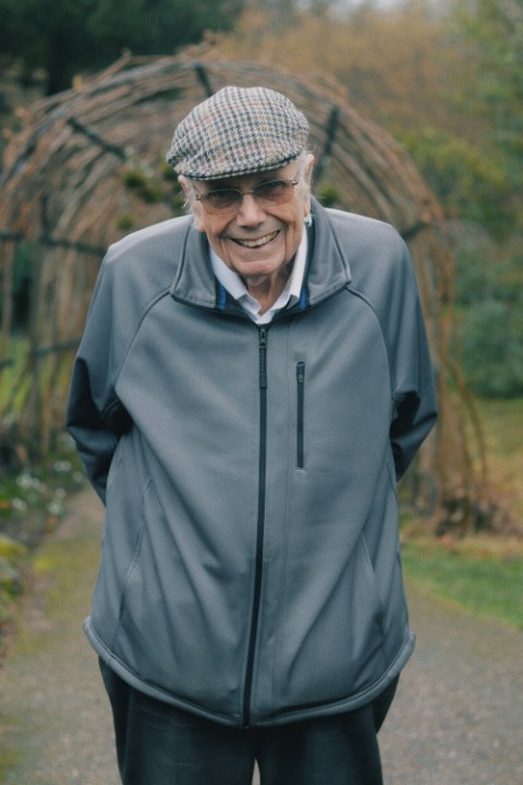 A warm portrait of a brightly smiling older gentleman standing in a quiet outdoor garden. He wears a tweed flat cap, glasses, and a gray jacket, with his hands comfortably behind his back.