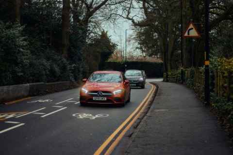 A vibrant red Mercedes-Benz drives toward the camera along a curving, tree-lined road. The sleek car and its bright headlights contrast sharply with the moody, overcast surroundings.