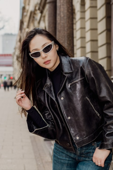 A chic woman posing on a city sidewalk. She wears a distressed black leather jacket, dark turtleneck, jeans, and striking white-framed sunglasses.