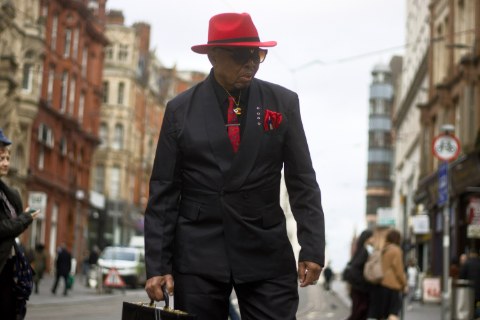 A stylish older gentleman walking confidently down a city street, wearing a black suit accented by a bright red fedora, tie, and pocket square, while carrying a briefcase.