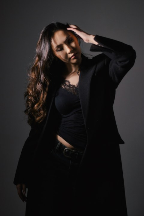 Dramatically lit portrait of a woman looking down, resting her hand in her long dark hair, wearing black.