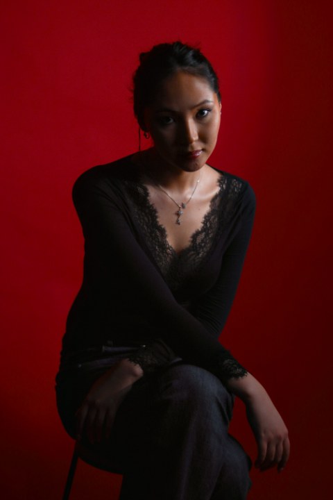 A moody portrait of a seated woman against a deep red backdrop. She wears a dark lace-trimmed top and delicate celestial jewelry.
