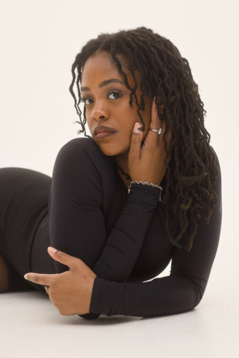 A close studio portrait of a woman with locs reclining on a white surface, wearing a black top and silver jewelry.