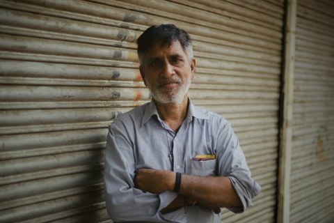 An older man with a graying beard stands outdoors with arms crossed. He wears a subtle striped shirt against a closed, textured metal shop shutter.