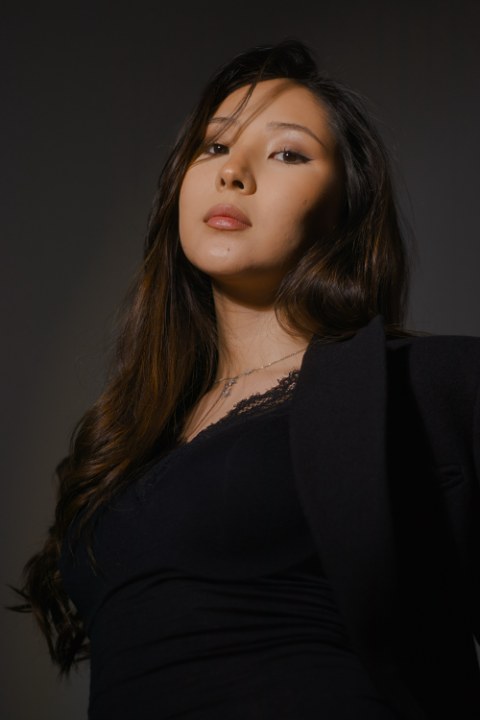 A dramatic, low-angle studio portrait of a woman looking downward toward the lens with a confident expression. She has long, dark, wavy hair and wears a black lace-trimmed top beneath a dark blazer, set against a deep charcoal backdrop.