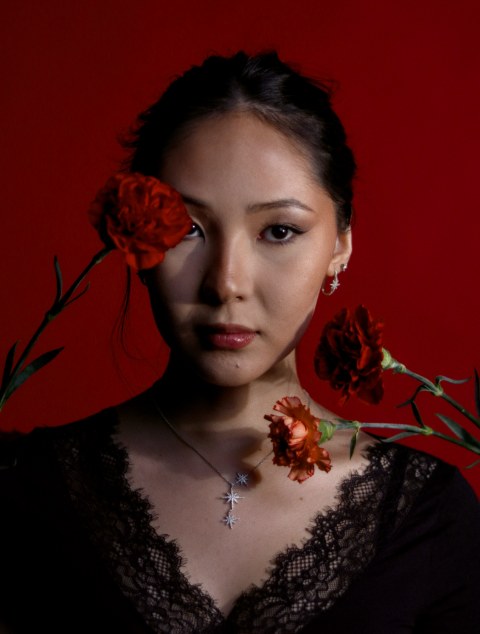 A dramatic portrait of a woman against a rich red backdrop, wearing black lace and star jewelry. Three crimson carnations cast sharp, artistic shadows across her face and neckline.