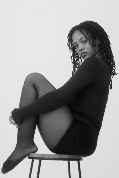A monochromatic studio portrait of a woman with locs, seated on a high stool with her knees tucked to her chest, embracing them. She looks directly at the camera over her left shoulder. She is wearing a black long-sleeved bodysuit and black sheer tights against a stark white, bright background.