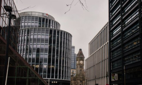 Modern curved glass offices surround an ornate, vintage clock tower in a city under an overcast sky.