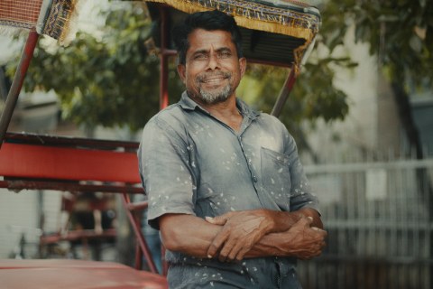 A portrait of a man with a graying beard standing outdoors, arms comfortably crossed, smiling warmly at the camera. He wears a paint-speckled blue button-down shirt and leans against a red cycle-rickshaw with a fringed canopy.