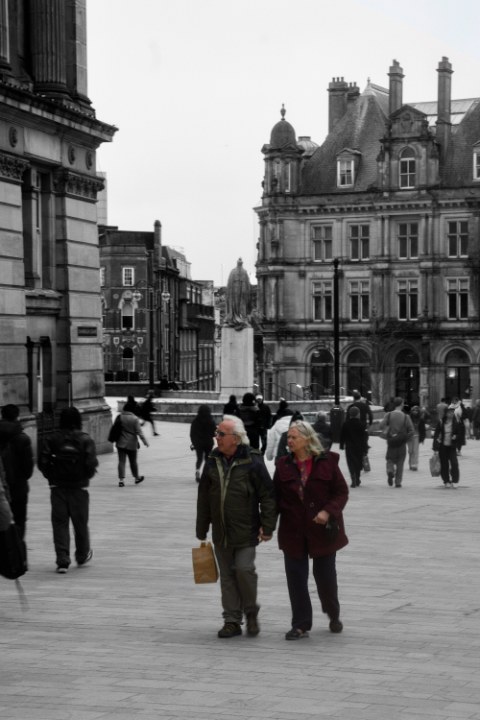 An older couple holding hands walks through a classic city square. The surrounding buildings are muted, subtly highlighting their presence together.