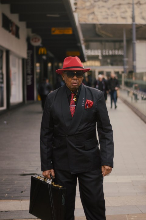A sharply dressed man walks outdoors holding a briefcase, wearing a dark suit, sunglasses, and a red fedora.